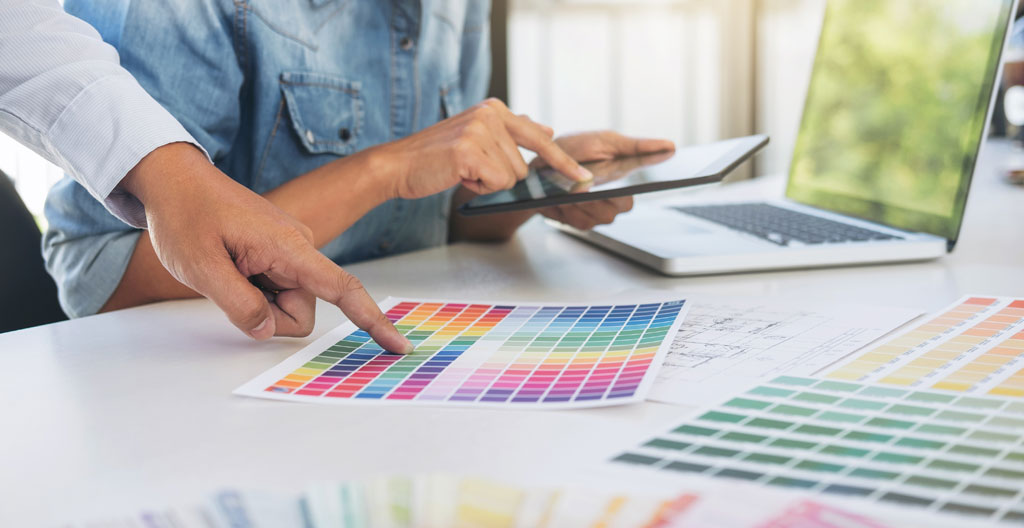 Vue de dessus d'une table de réunion d'agence graphique avec planches de tendances, échantillons de papiers, maquettes imprimées, iPad affichant wireframes, mains professionnelles annotant les documents lors d'une session de travail collaborative
