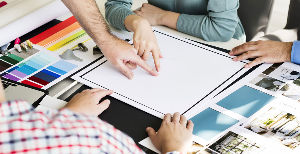 Progression de création de logo sur table de travail de graphiste, de gauche à droite : croquis au crayon, vectorisation sur tablette graphique avec écran, impression test couleur annotée, entourés d'outils professionnels sur surface bois clair